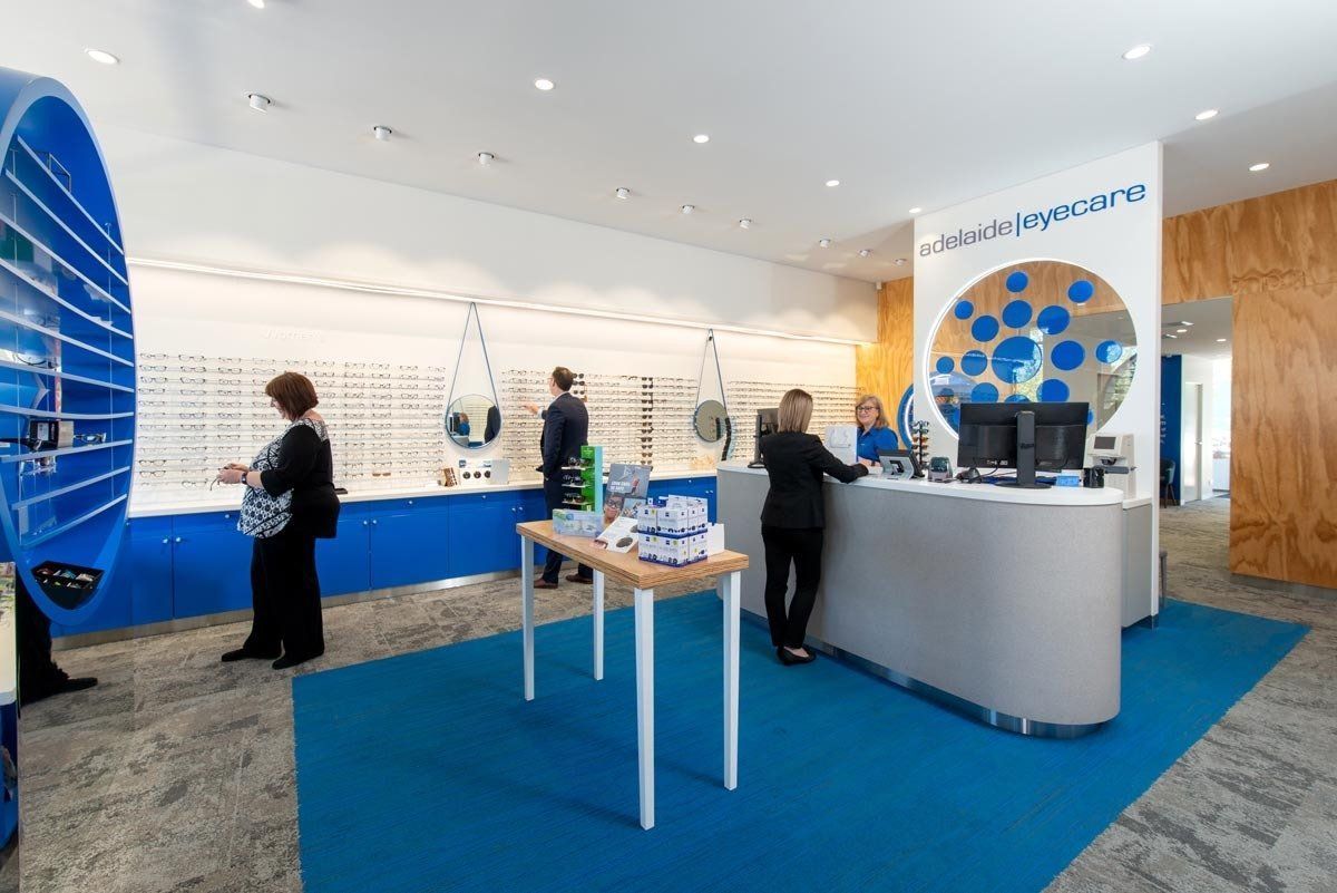 An optical store interior with customers browsing eyeglasses. Blue accents, reception desk, and displays.