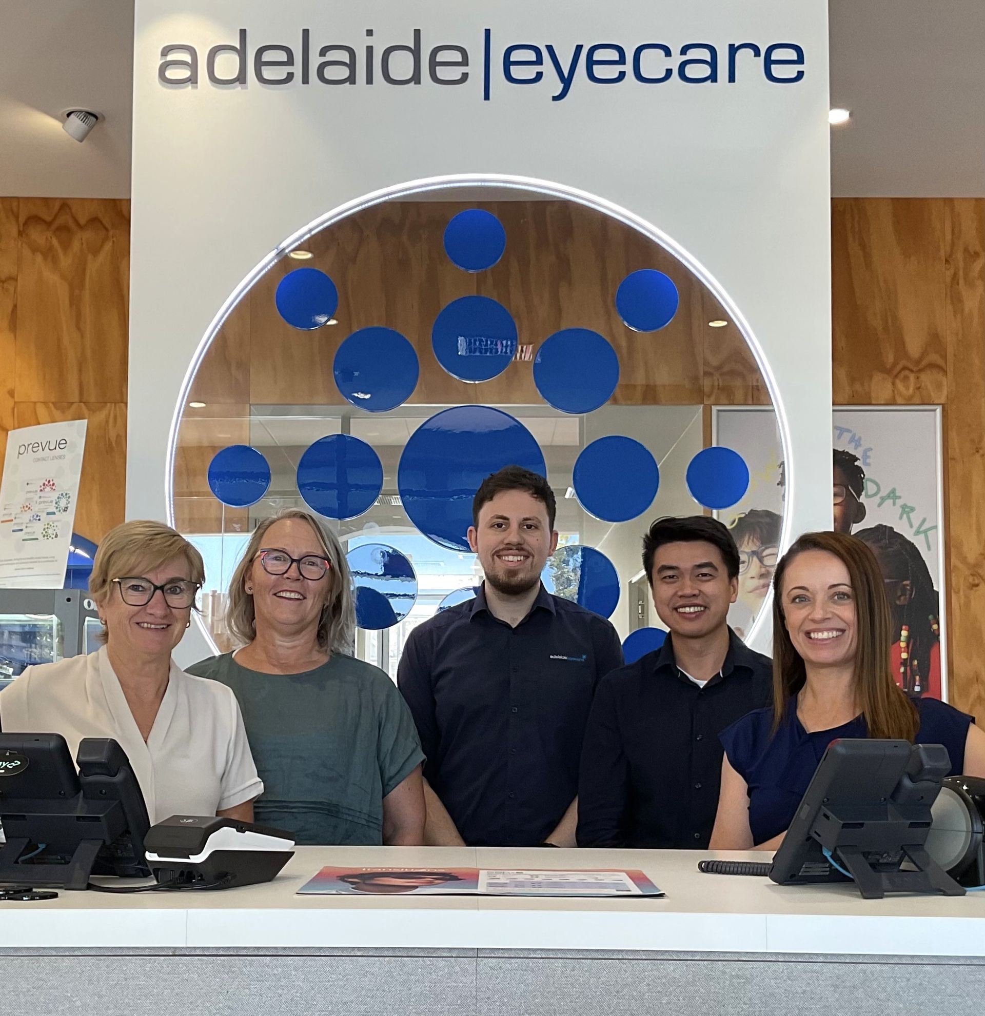 Adelaide Eyecare staff pose behind a counter. The sign above has blue circles. The team members are smiling.