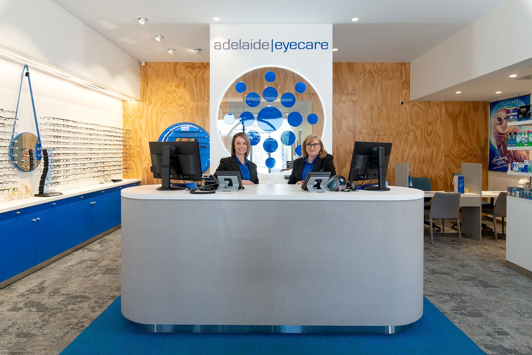 Two people at a modern eye care reception desk. Blue and wood accents.